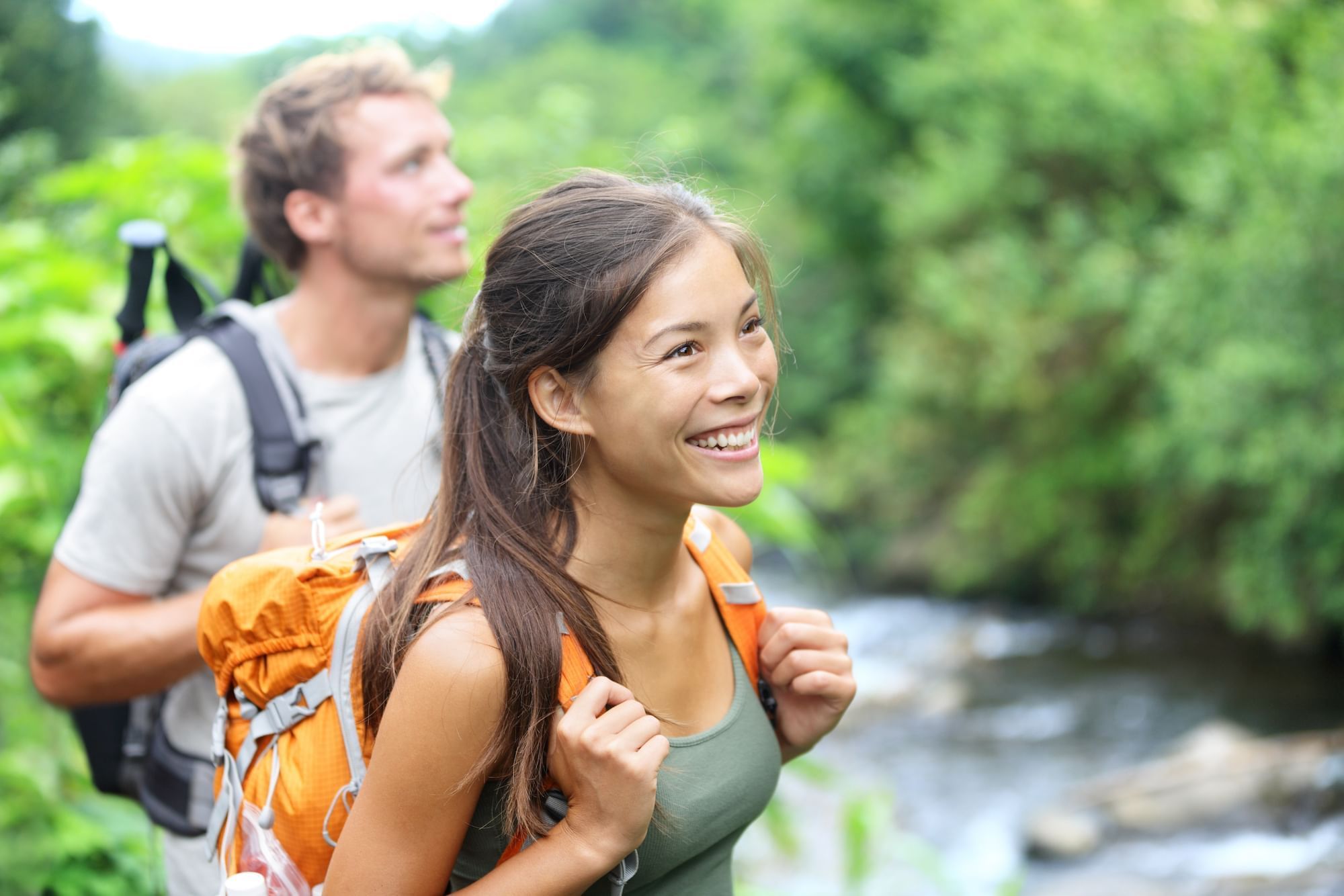 Smiling couple with backpacks hiking along a stream surrounded by lush greenery near Cove Pocono Resorts