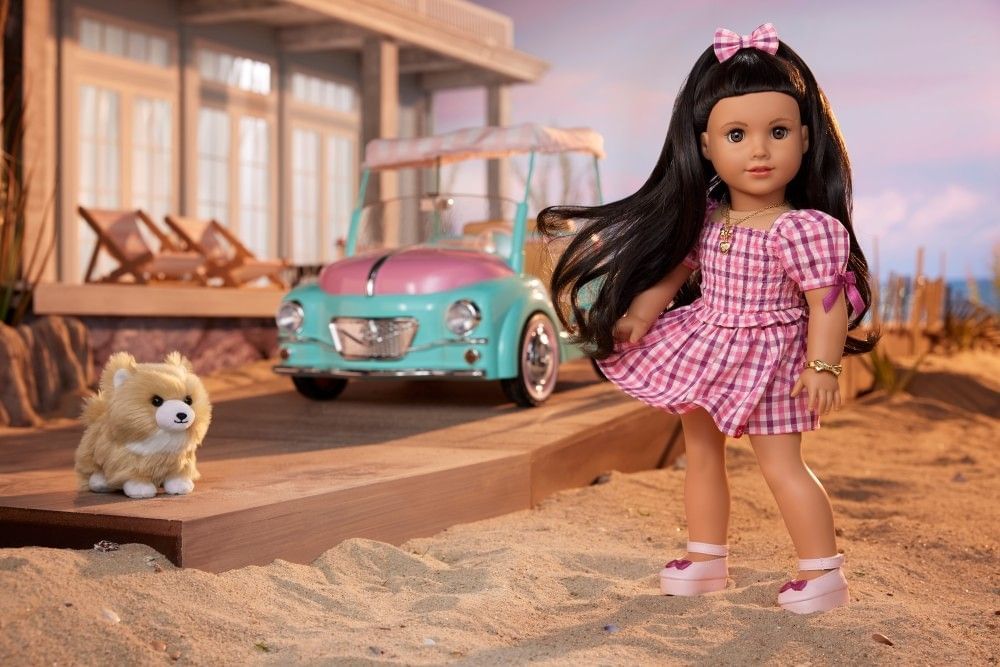 A doll with brown hair beside a toy car and dog on a sandy beach.