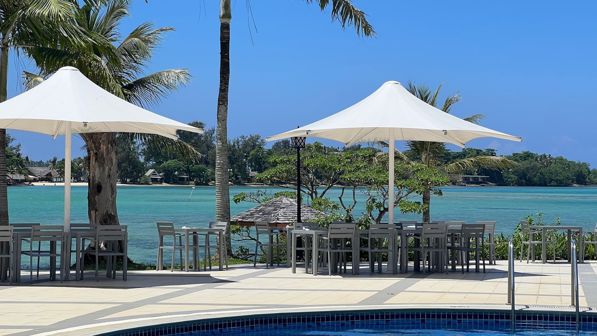 Pool Grill & Bar area with beach view & white umbrellas by a blue pool overlooking a turquoise bay at Warwick Le Lagon Efate