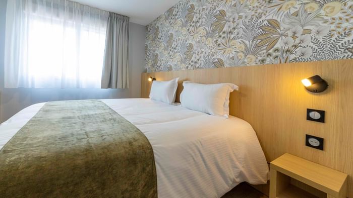 A cozy hotel room with a white bed, wooden headboard, and nightstand with lamps at The Originals City Hotel du Parc Gravelines.