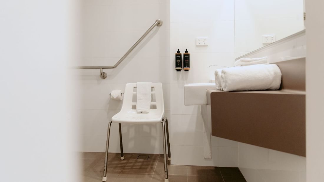 A bathroom with a chair, sink, mirror, and two bottles on a shelf.