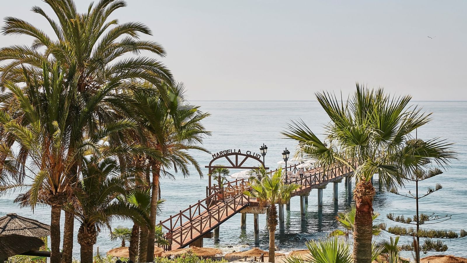 Wooden pier with the's name extending over the calm blue sea, framed by several palm trees at Marbella Club