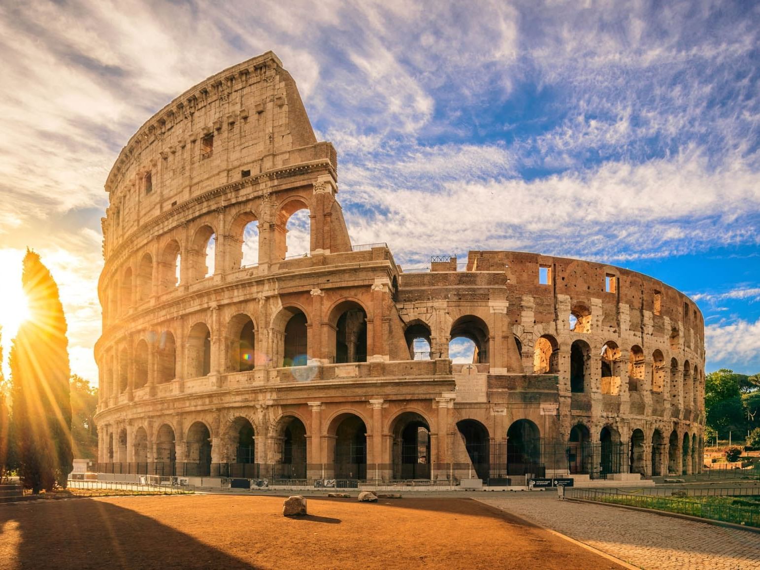 Sunlit Colosseum in Rome with ancient arches beneath a vivid blue sky and wispy clouds, near The Guardian Hotel
