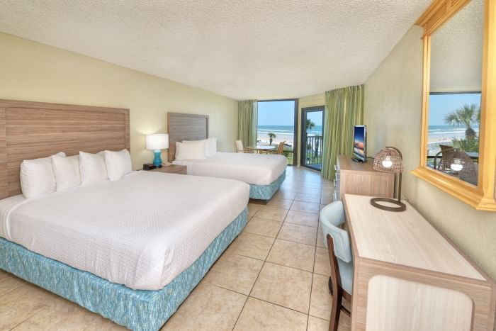 A wide-angle view of a hotel room with two queen beds, a desk, and a balcony with a beach view.