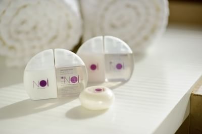 Amenities de marca junto a toallas enrolladas bajo luz suave sobre superficie blanca en NOI Vitacura