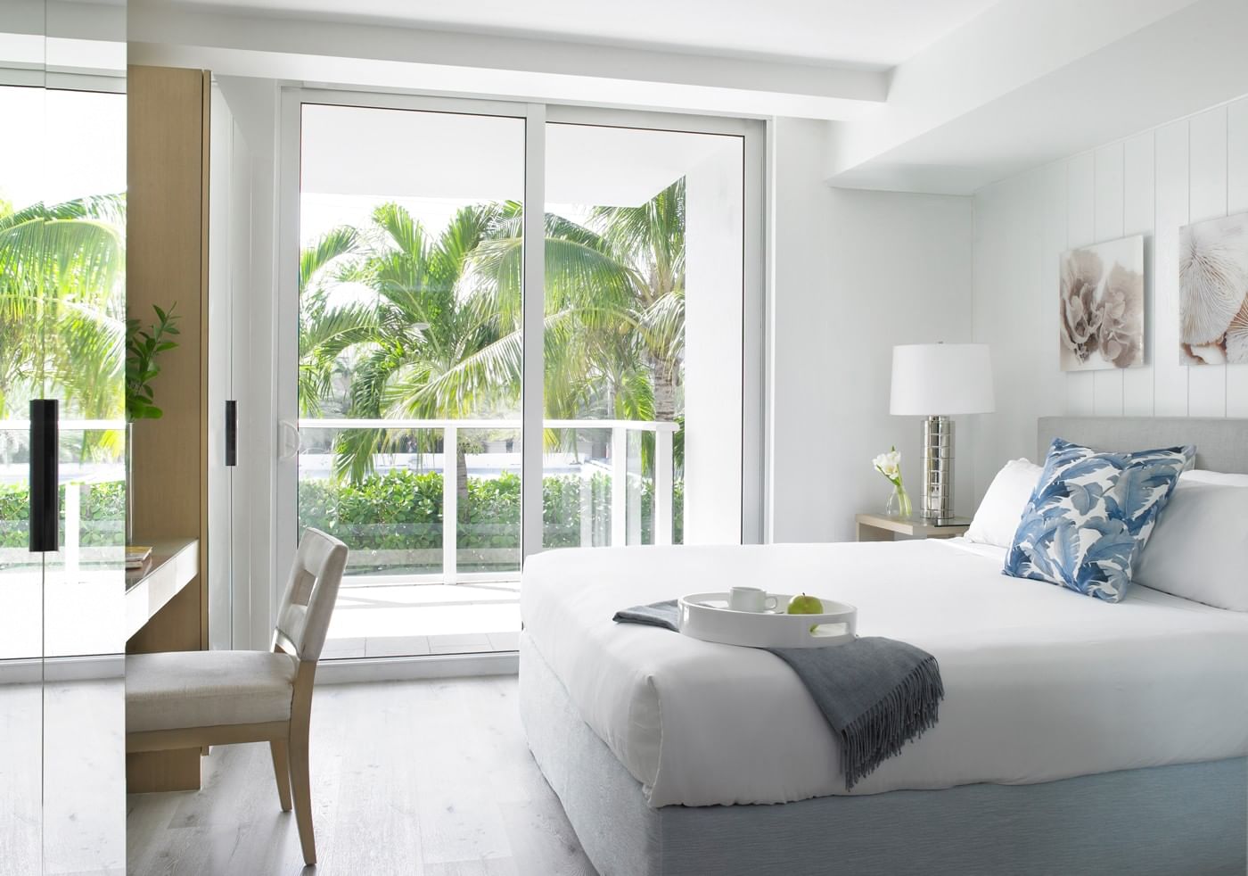 A bright and airy hotel room with a large bed, desk, and balcony overlooking palm trees.