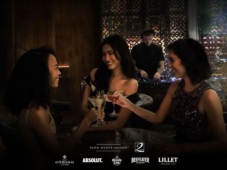 Three women toasting with cocktails as a DJ plays in the background in 2 Lam Son at Park Hyatt Saigon
