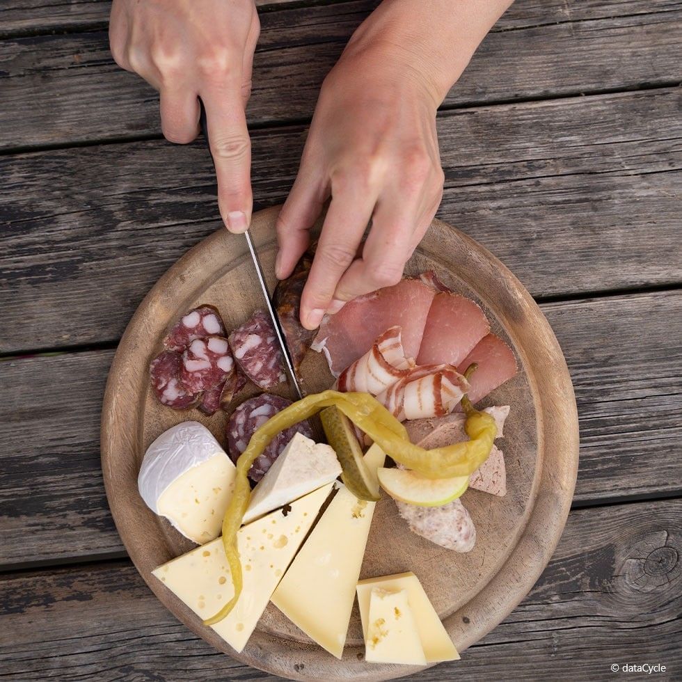 Hands cutting a piece of salami on a wooden plate with assorted meats and cheeses.