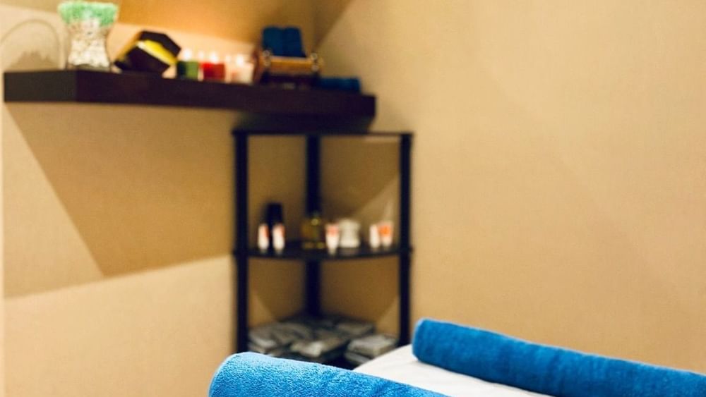 Spa room, featuring blue rolled towels on a bed near candles and oils by a corner shelf at Naviti By Warwick Dammam