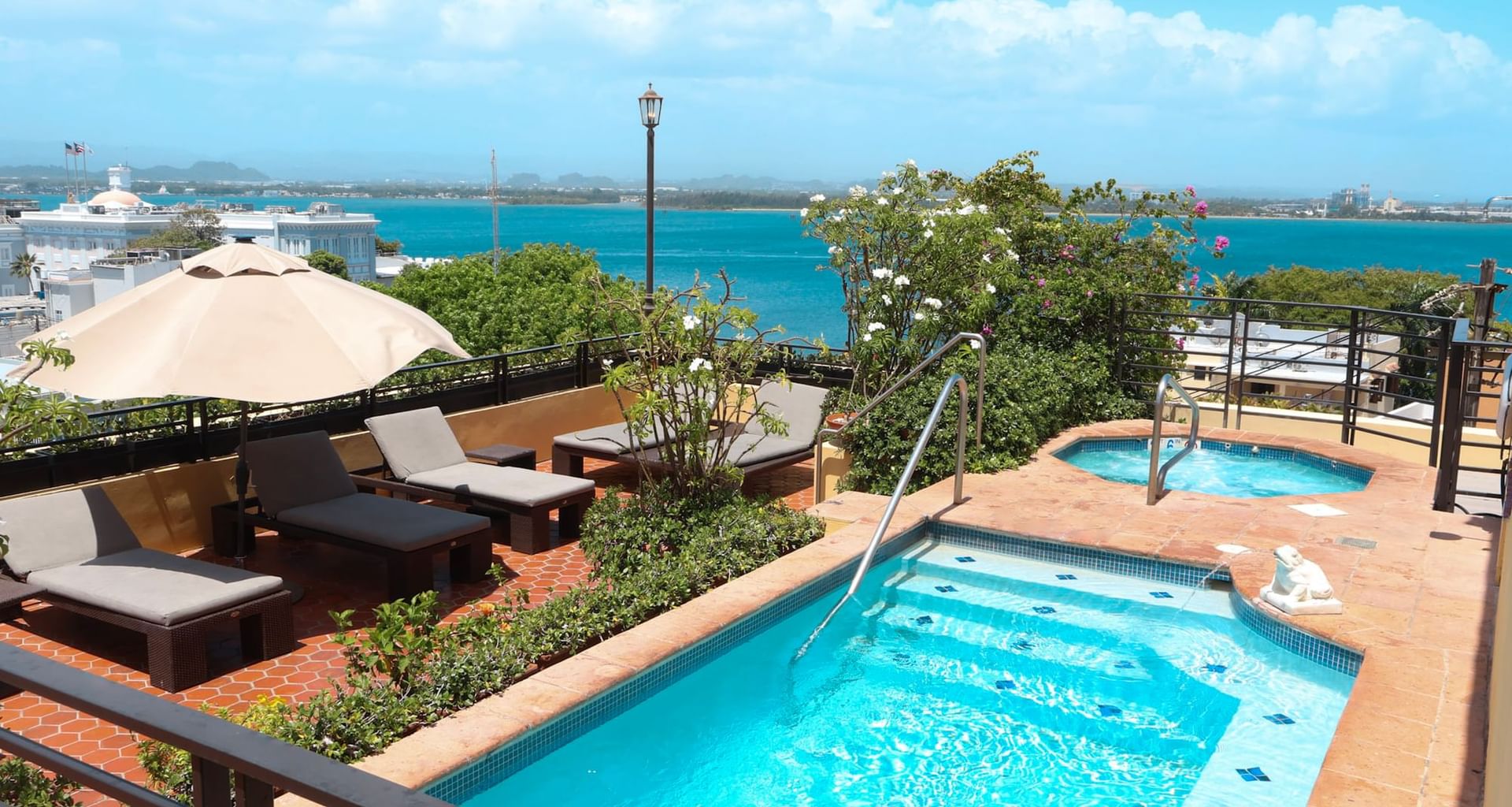 Outdoor rooftop area with two pools, lounge chairs, umbrella, and ocean view in the background.