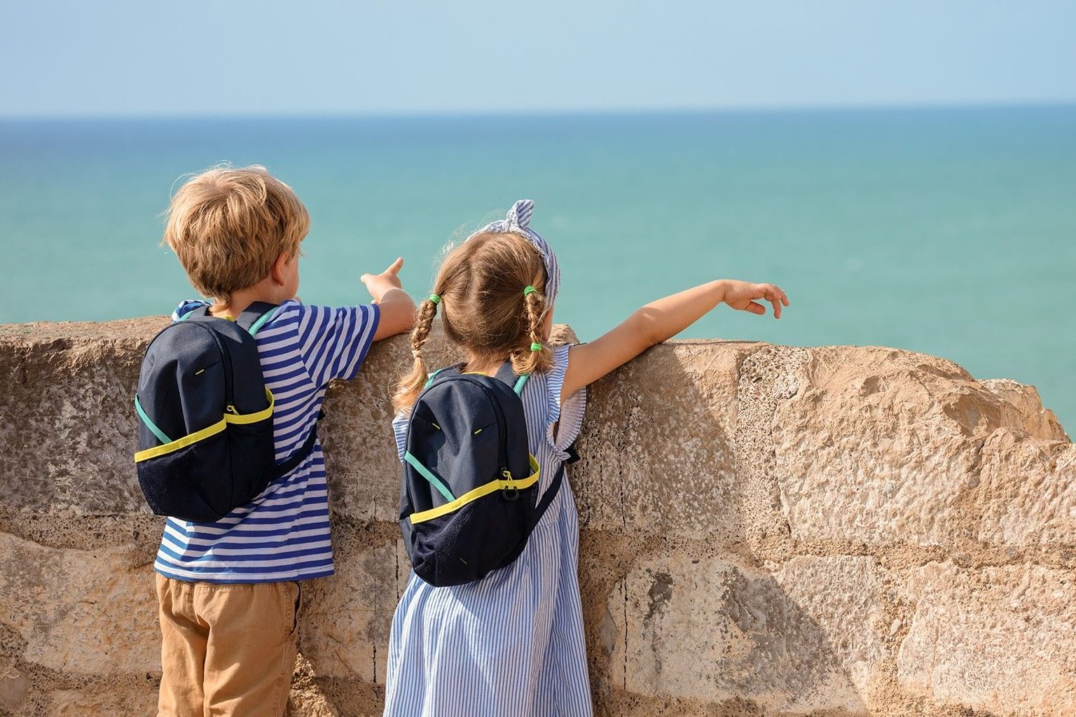 Two young children with small black backpacks stand at a stone wall, looking out at the turquoise ocean on a sunny day.