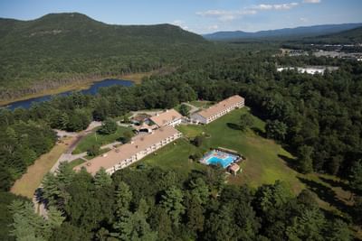 Aerial view of the Fox Ridge Resort buildings, pool, and lush forest with mountains and a lake in the background.