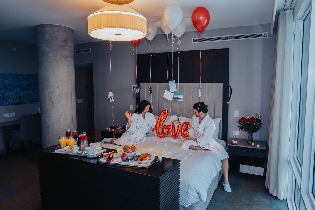 Couple sitting on a bed with a love balloon and breakfast tray, under white and red balloons at Hotel X Toronto