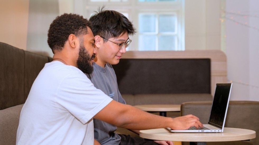 Two men sit at a table, one using a laptop while the other watches.