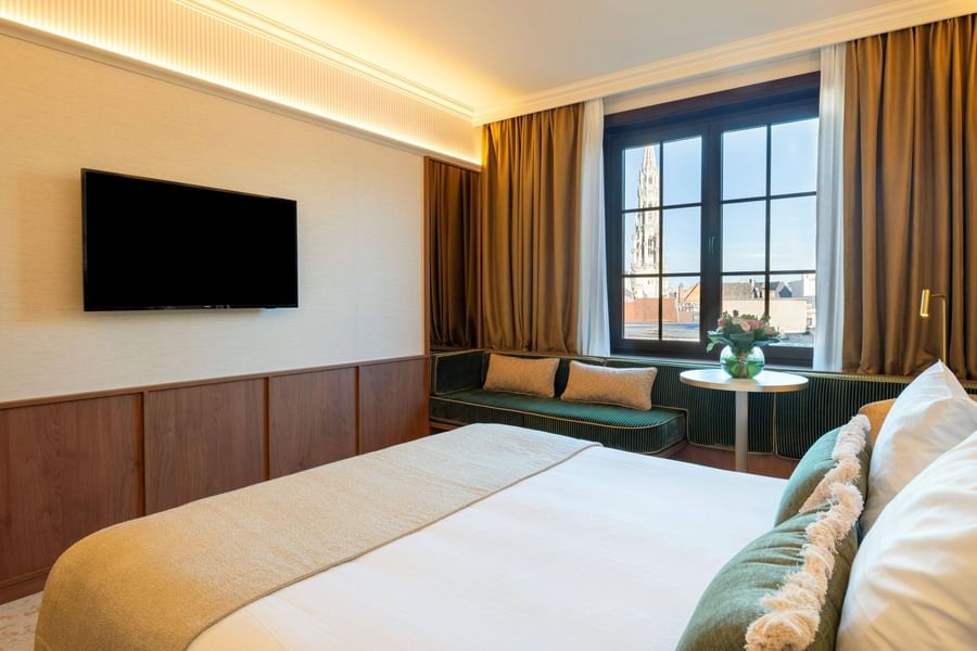 Modern hotel room with white bed, wall-mounted TV, window seating area, and city views at Warwick Brussels Grand-Place