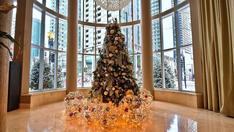 Decorated Christmas tree with ornaments and lights in a lobby with large windows and urban view.