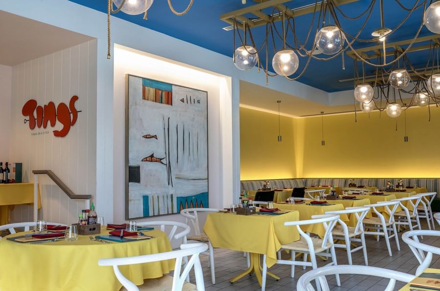 Yellow tables by white chairs under globe lights in Tings on a Stick painting at Warwick Paradise Island Bahamas