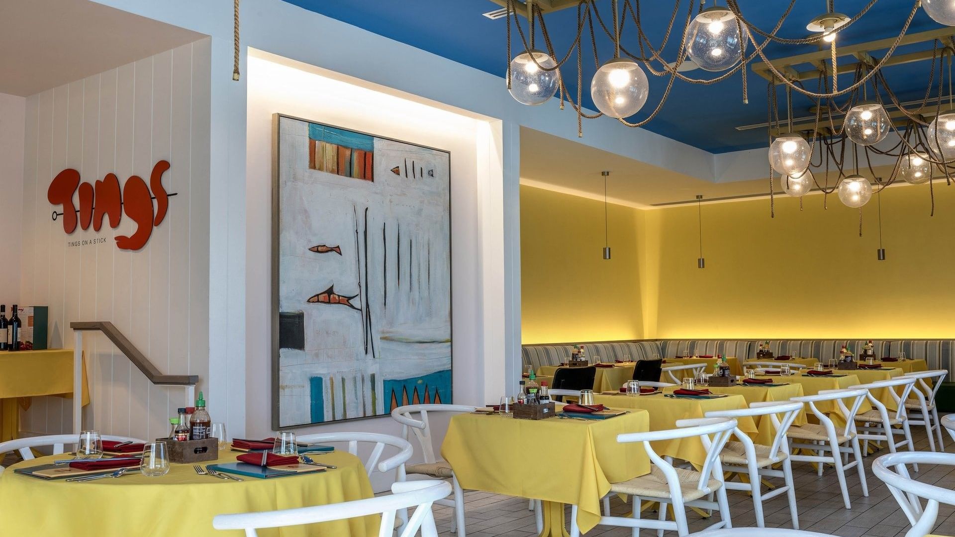 Yellow tables by white chairs under globe lights in Tings on a Stick painting at Warwick Paradise Island Bahamas