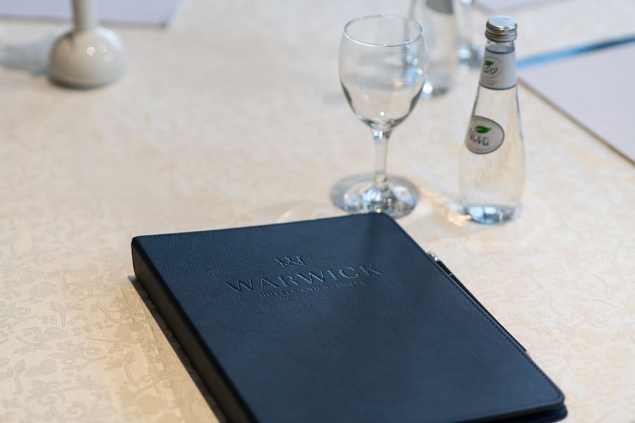 Warwick logo on a leather folder by a glass and water bottle on a white table in Amarige Hall at Warwick Hotels and Resorts