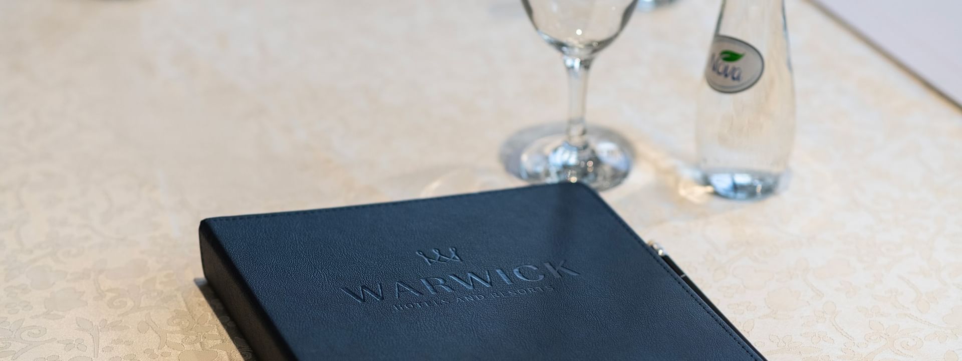Warwick logo on a leather folder by a glass and water bottle on a white table in Amarige Hall at Warwick Hotels and Resorts