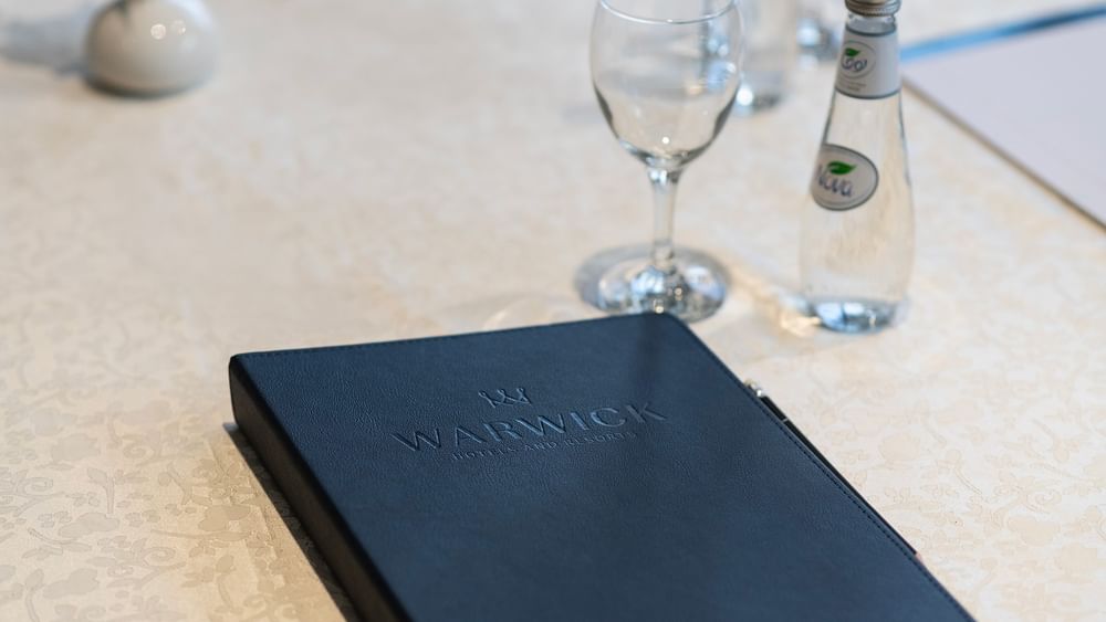 Warwick logo on a leather folder by a glass and water bottle on a white table in Amarige Hall at Warwick Hotels and Resorts