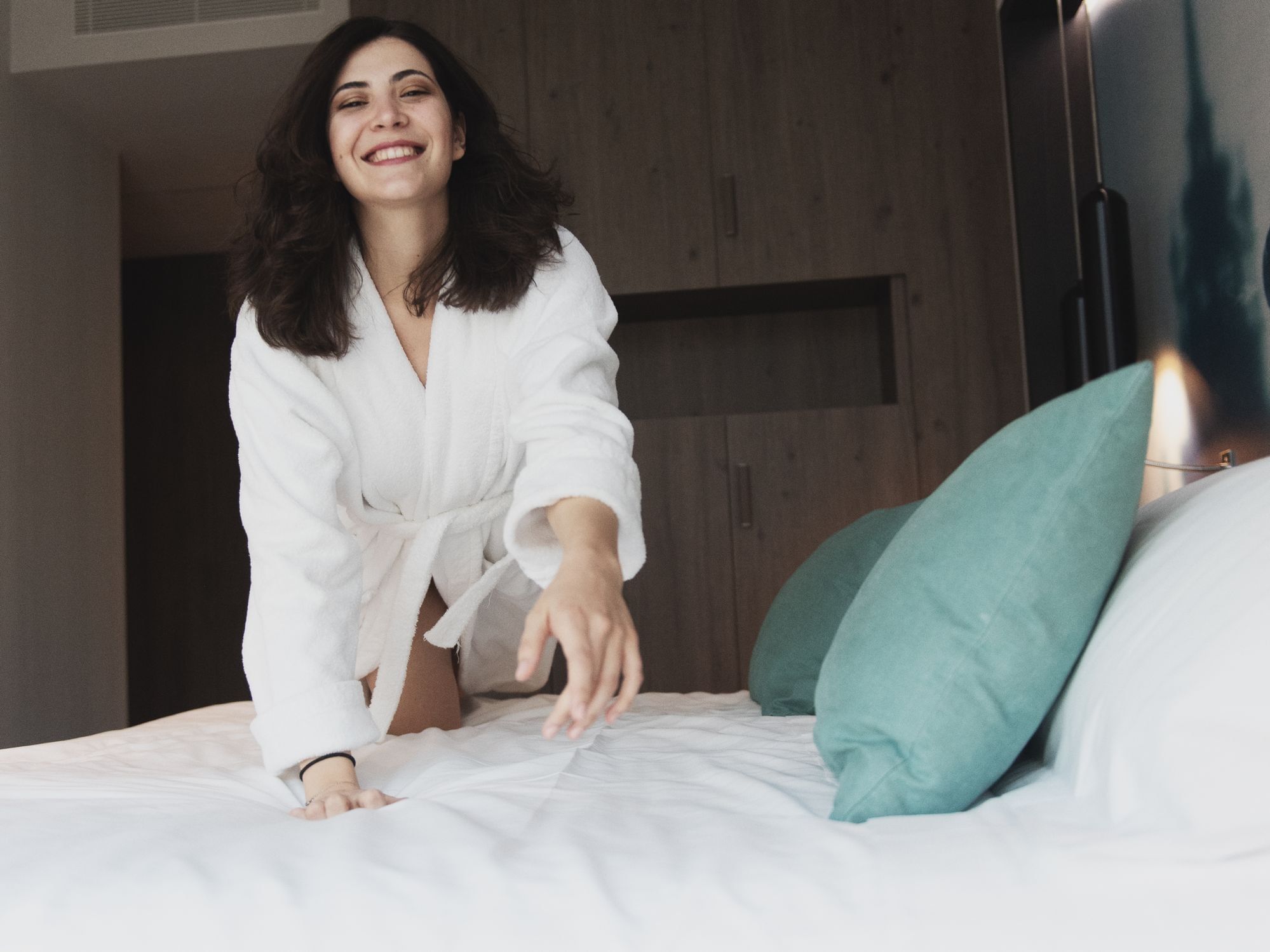 Woman in bathrobe reaching for bed with green pillows, evoking relaxation and comfort.