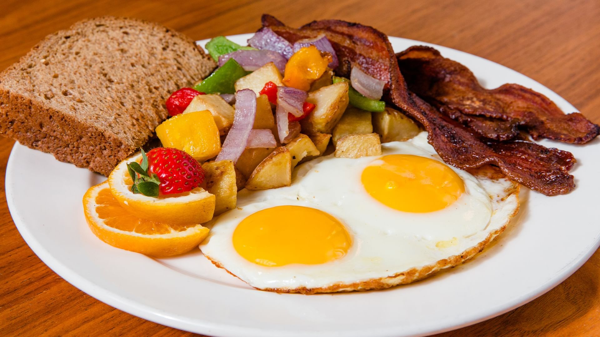A plate of breakfast food with eggs, bacon, toast, fruit, and potatoes.
