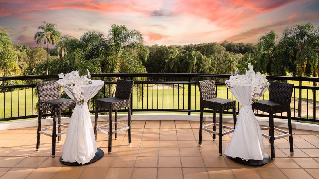 Two tables with white cloth and flowers on a balcony overlooking a golf course with palm trees at sunset.