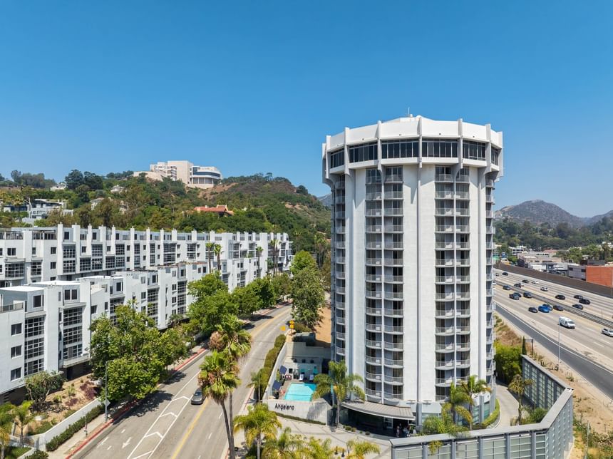 Wide aerial shot of the circular Hotel Angeleno tower next to the freeway, with hills and the pool area visible