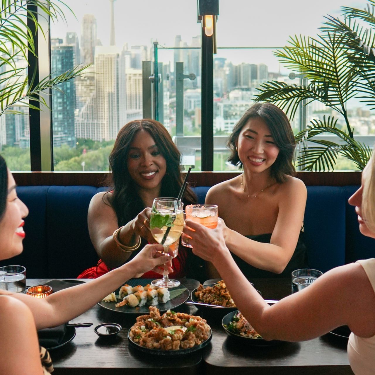 Four women are dining and toasting with drinks at a table with a city skyline view.