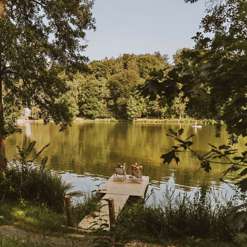 Zwei Personen sitzen auf Liegestühlen auf einem Steg an einem ruhigen See.