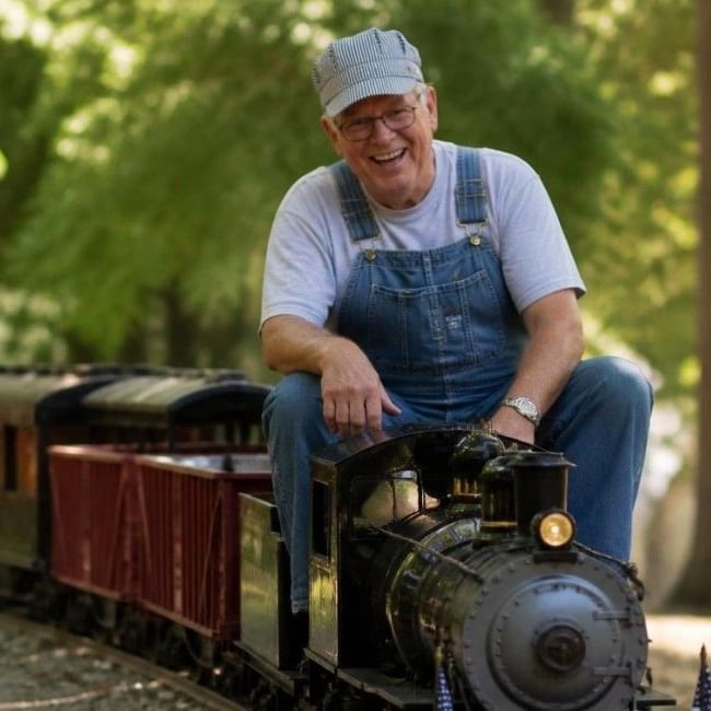 A smiling volunteer in dungarees and cap rides a miniature steam locomotive through a wooded setting at Pinewood Miniature Railway