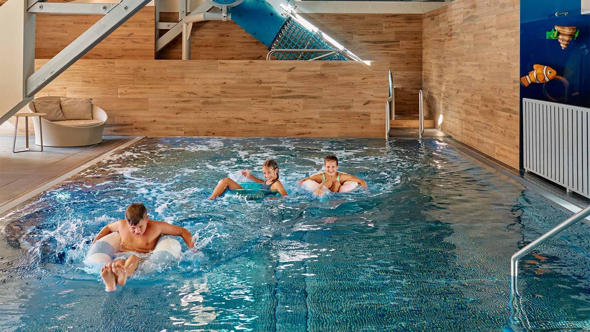 Three kids are playing in an indoor pool with a slide and a chair nearby.