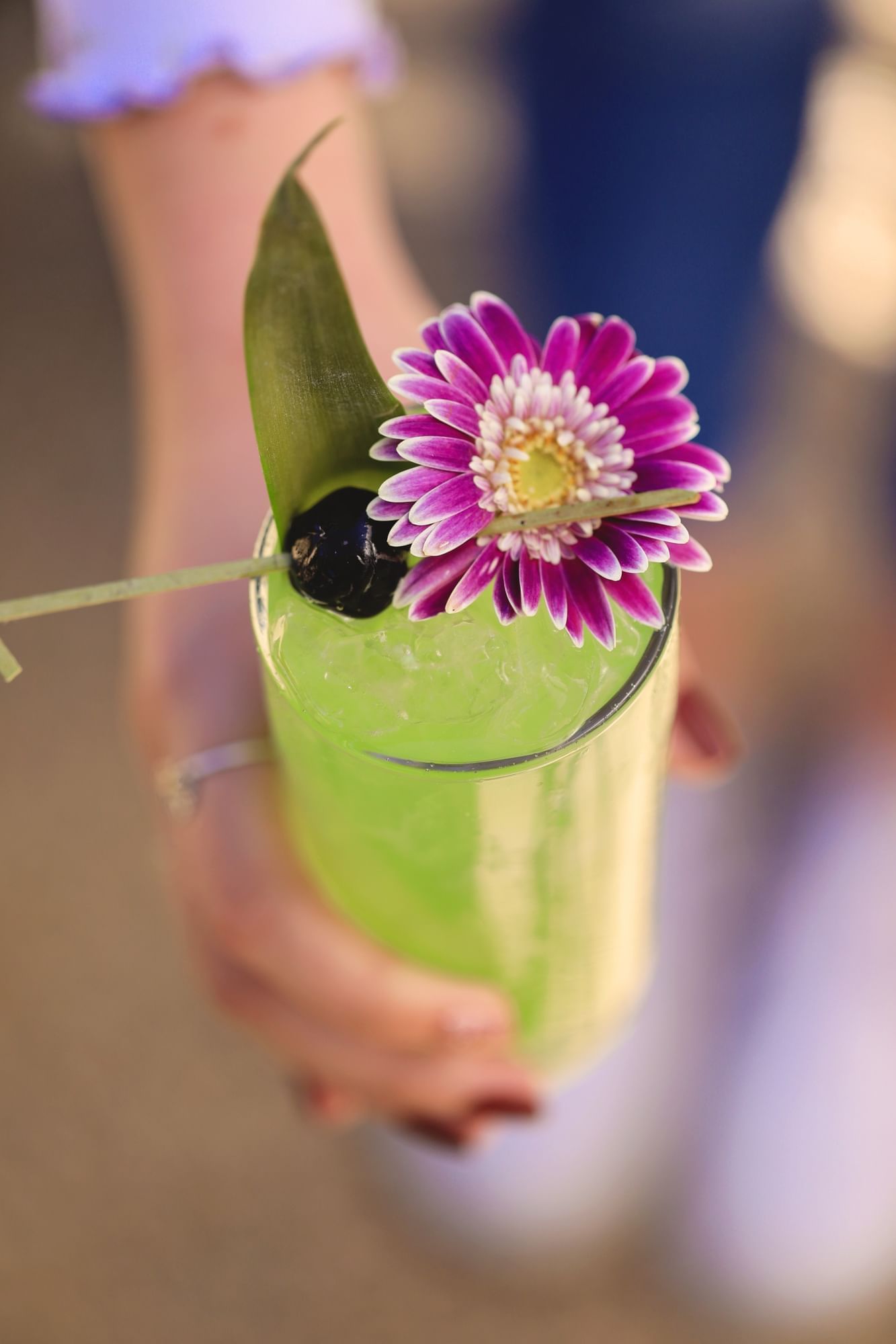 Lorem ipsum A person's hand holding a clear glass filled with a bright green cocktail and a purple flower on top