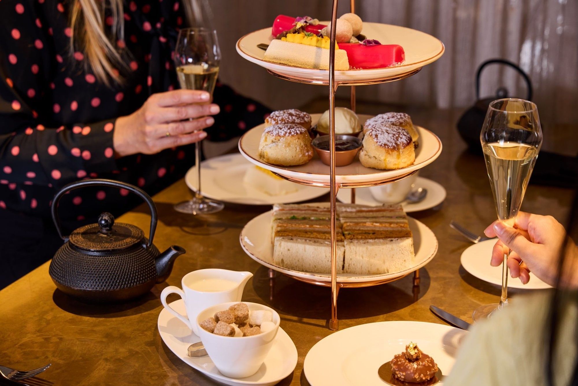 People enjoying a three-tiered assortment of cakes, scones and sandwiches with champagne in May Fair Kitchen
