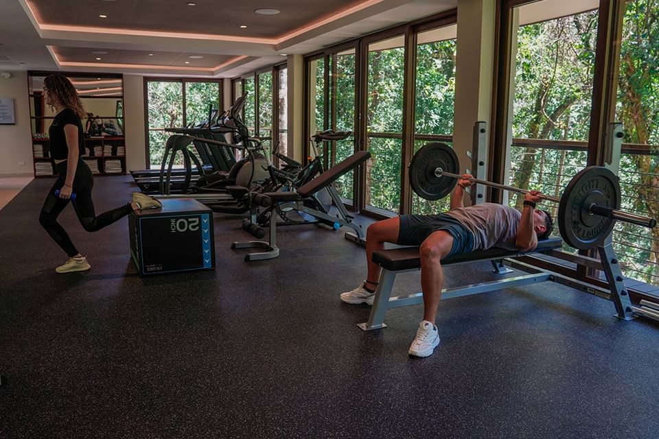 Two people exercising in a gym with large windows showing trees at El Silencio Lodge & Spa