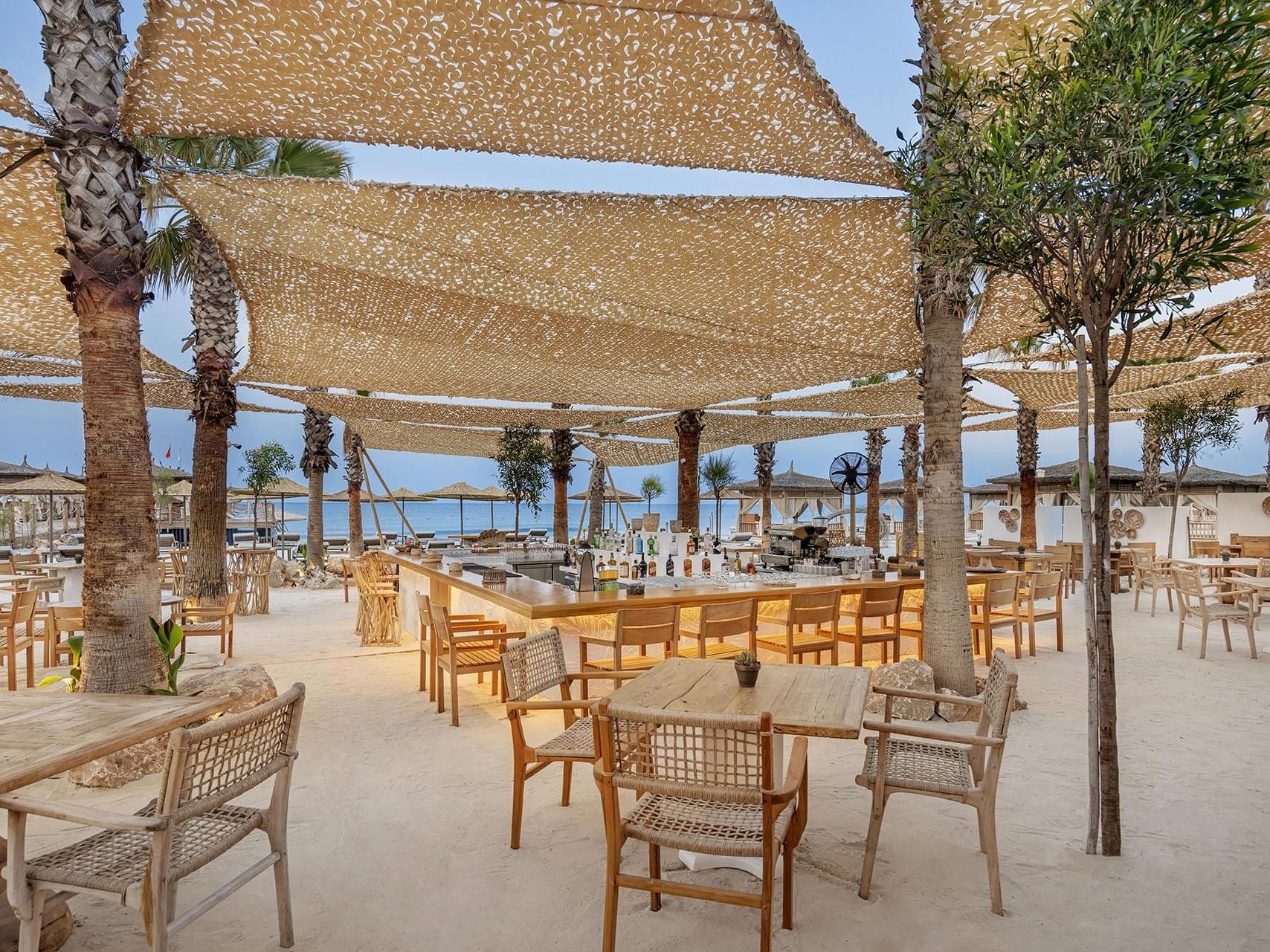Cullinan Belek: Outdoor beachside bar with wooden tables, chairs, and a canopy, surrounded by palm trees and sand.