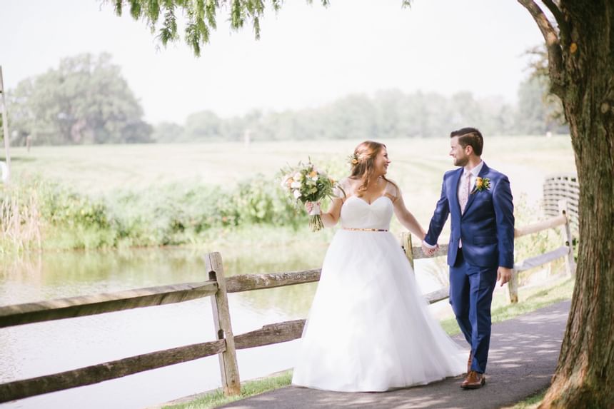 Bride in white gown and groom in blue suit hold hands by wooden fence near pond at Aramark Chauncey Conference Center