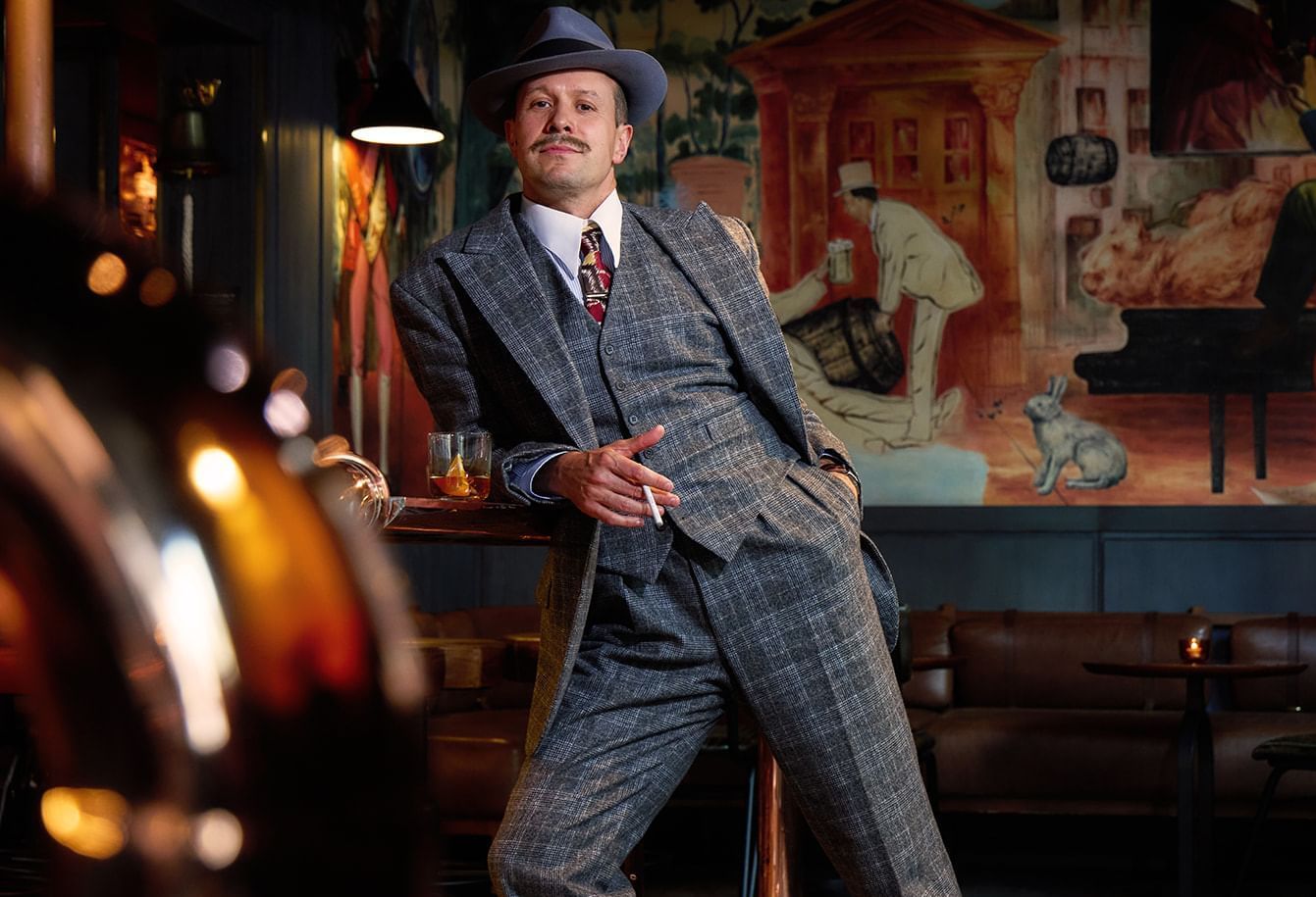A dapper man in a vintage suit and fedora posing confidently in The Midnight Bell bar at The Londoner Hotel