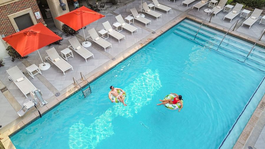 Two guests on leaf-patterned floats by a ladder in a bright blue swimming pool at Warwick Melrose Dallas