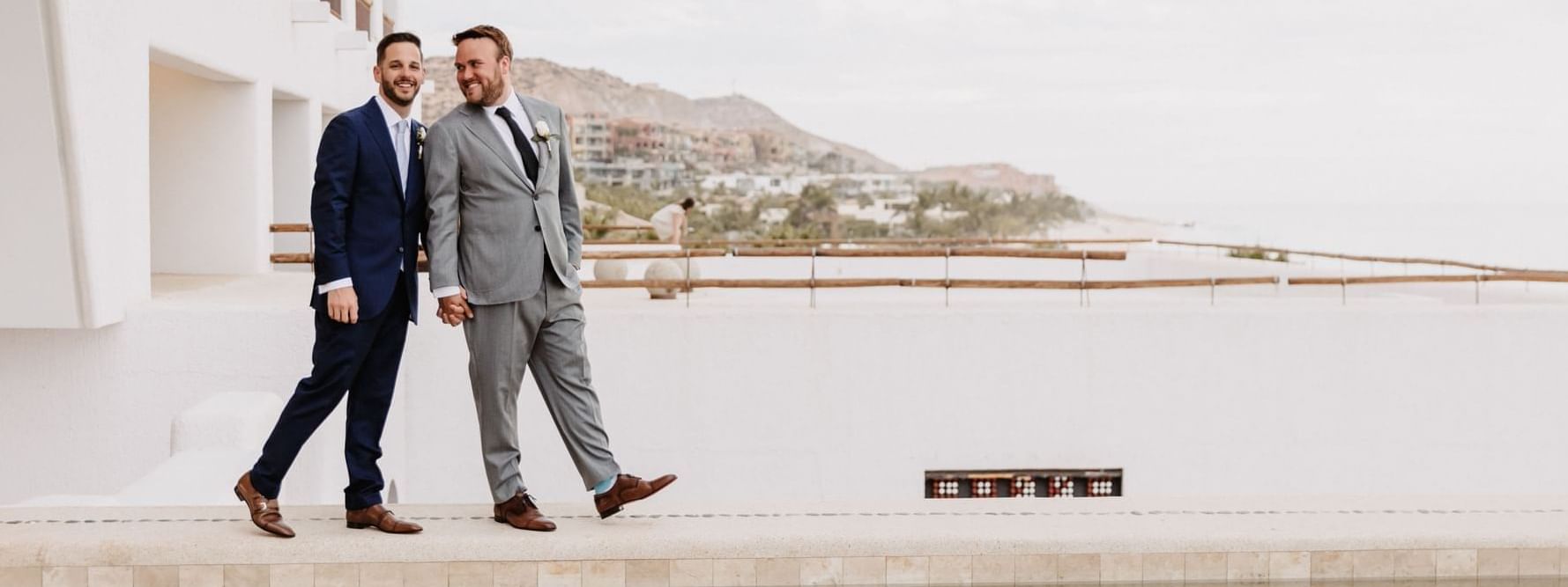 Two grooms holding hands and smiling on a rooftop patio at the Marquis Los Cabos Resort