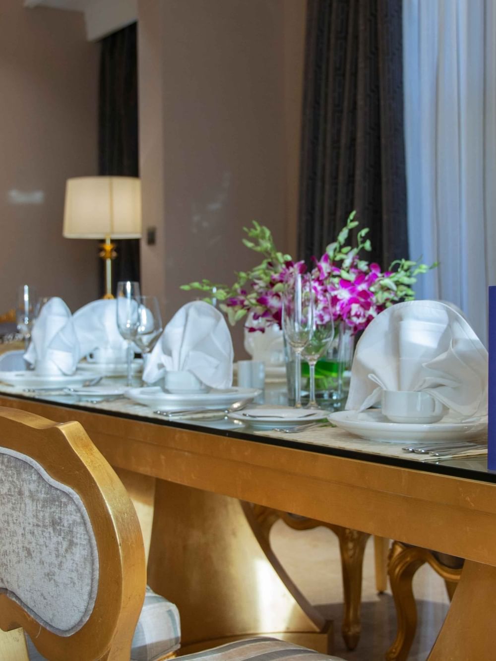 Gilded dining chairs by a glass-top table featuring fresh orchids & fine dinnerware in Penthouse Villa at Warwick Al Khobar