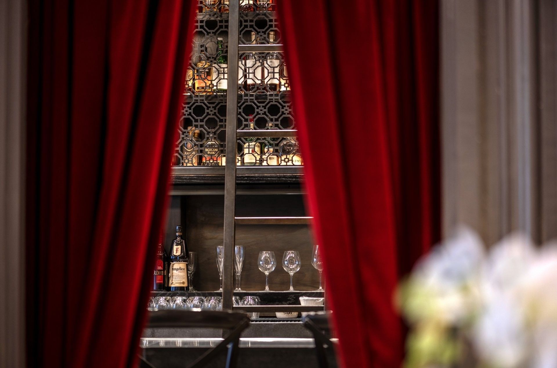 Liquor bottles on a backlit rack by wine glasses under red curtains in The European at Warwick San Francisco