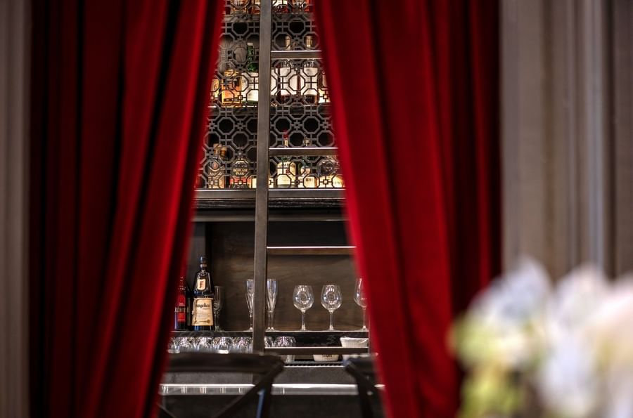 Liquor bottles on a backlit rack by wine glasses under red curtains in The European at Warwick San Francisco