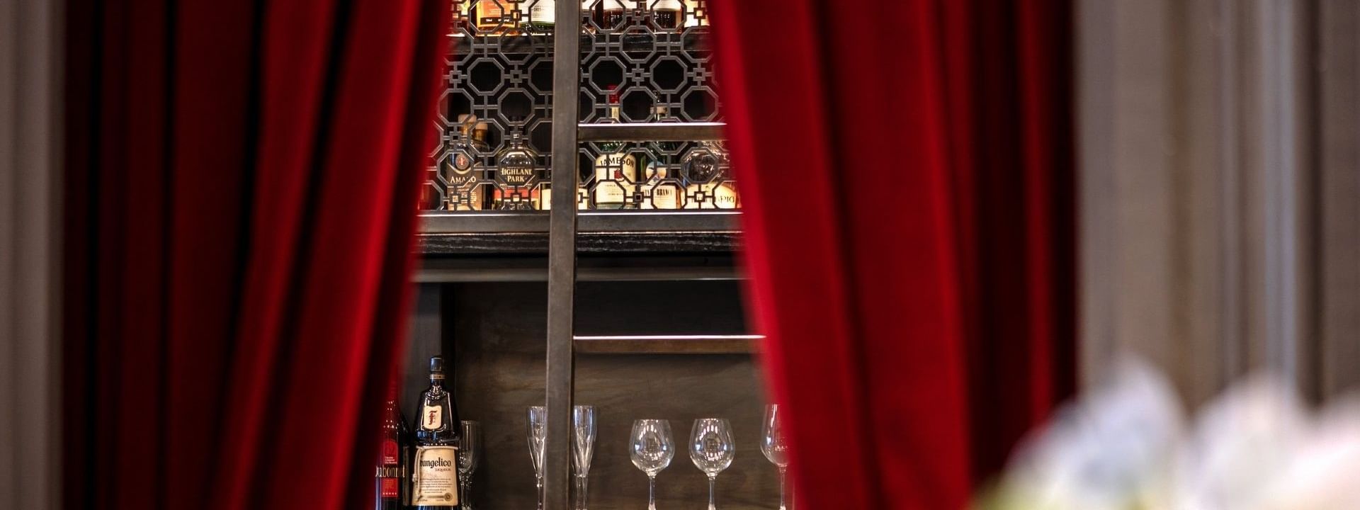 Liquor bottles on a backlit rack by wine glasses under red curtains in The European at Warwick San Francisco