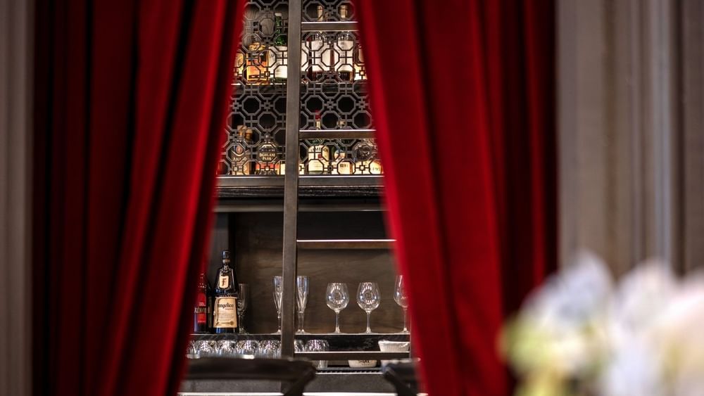 Liquor bottles on a backlit rack by wine glasses under red curtains in The European at Warwick San Francisco