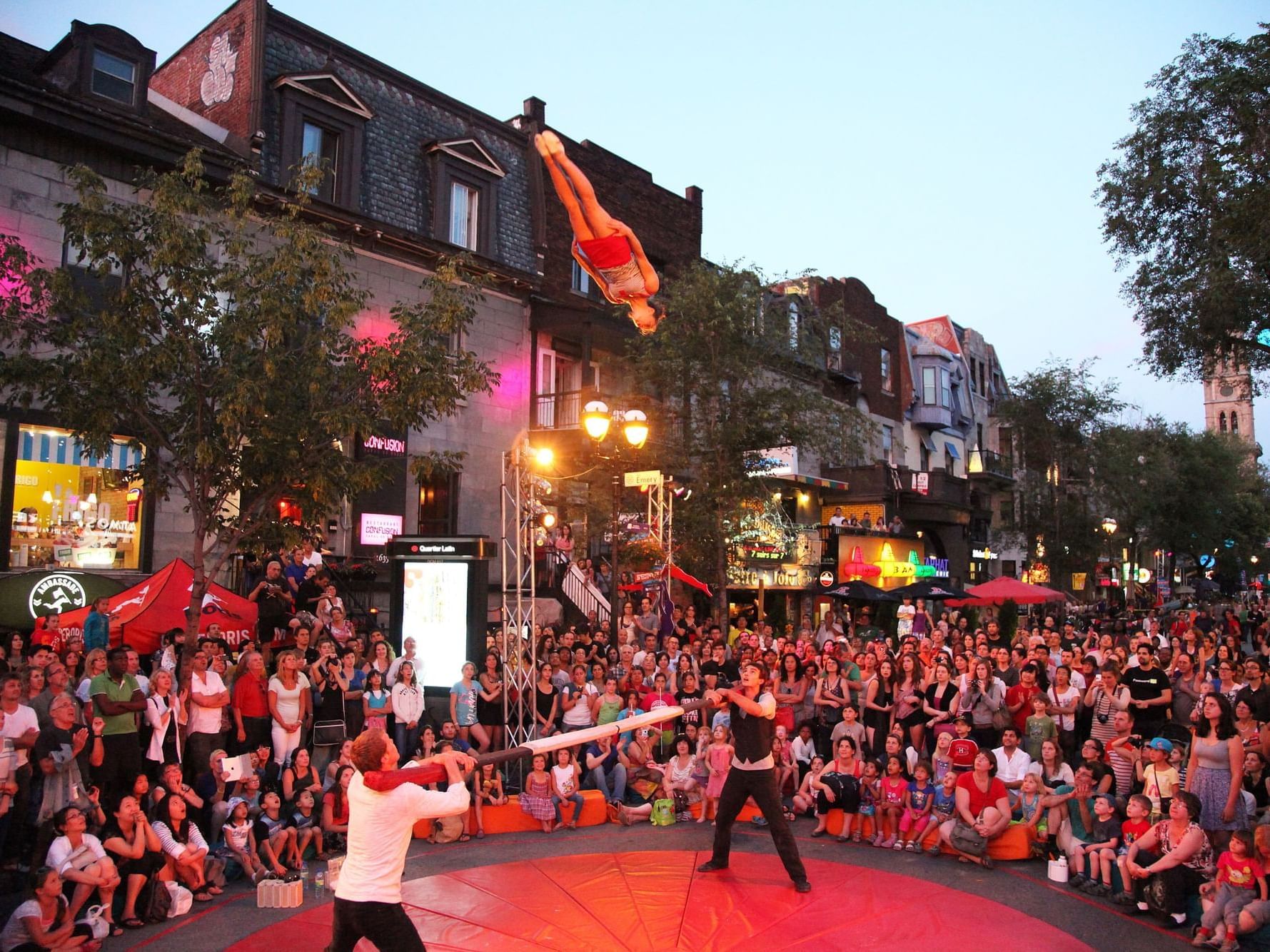Performers doing acrobatics with a large rod in front of a crowd in an outdoor city setting.