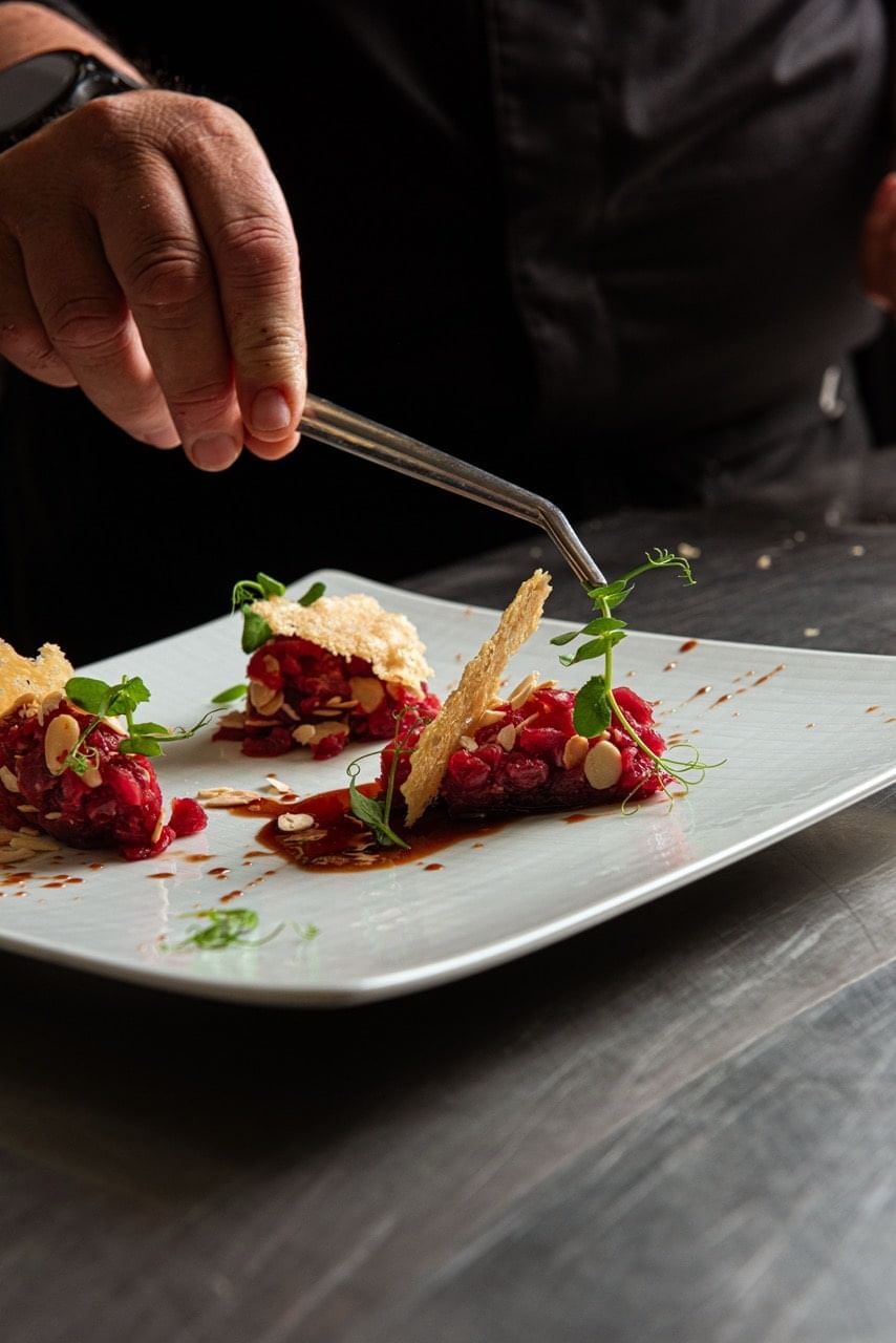 A hand garnishes a colorful dessert with a leaf, fruit, crunch, and sauce at Diana's Place, AG Hotels