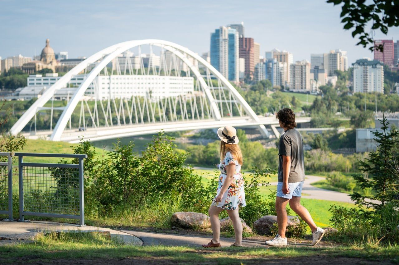 Couple walks along a park pathway near a bridge and cityscape in the background.