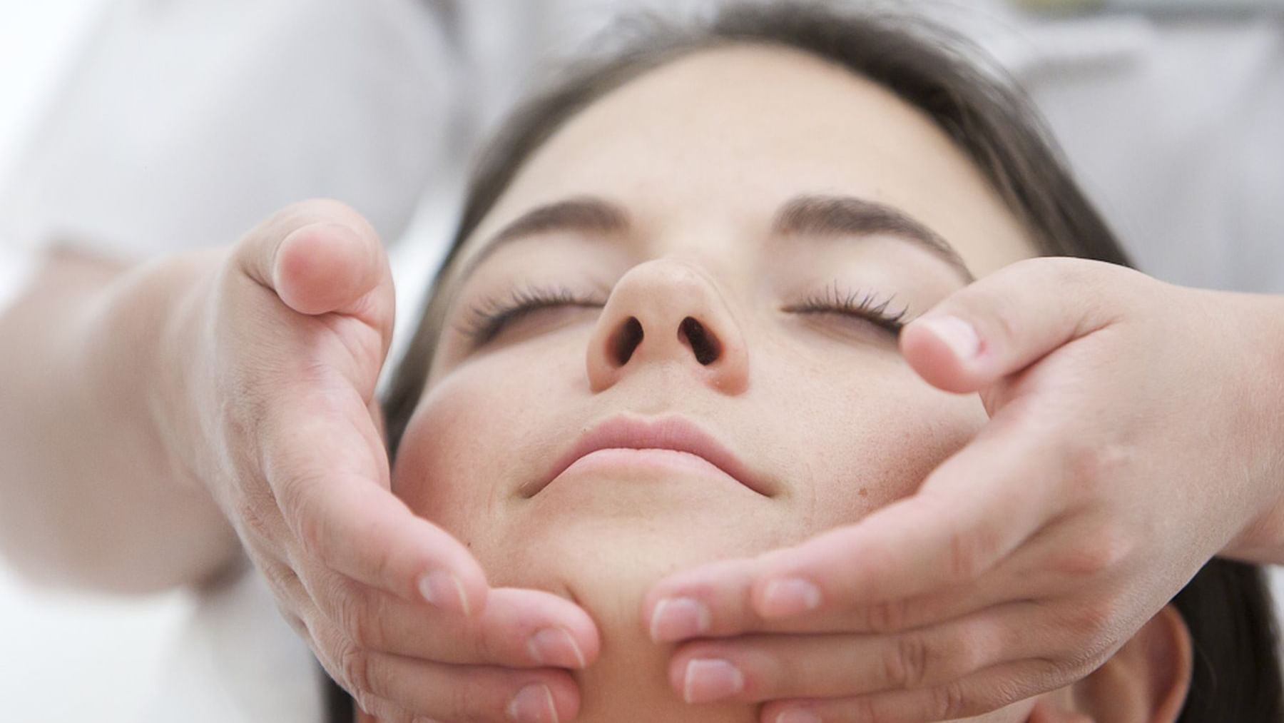 A woman receives a facial massage with hands on her forehead and cheeks, eyes closed.
